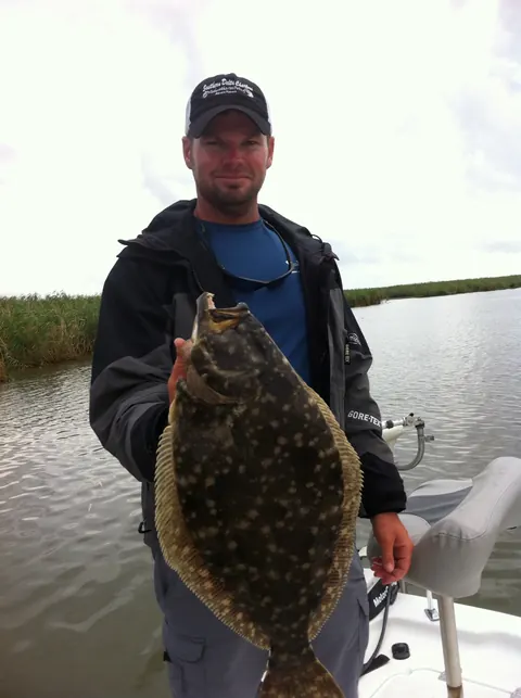 Angler in a dark jacket holding a flounder with marsh grass behind him near Venice, Louisiana