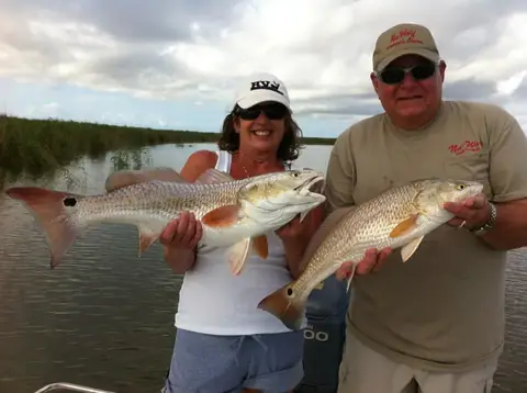 Two anglers each holding a redfish in front of delta marsh grass